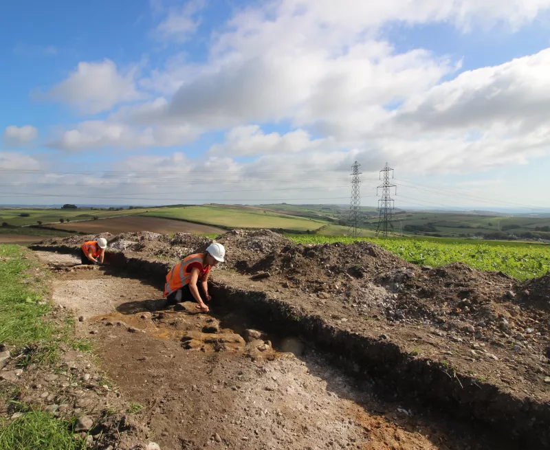 Image showing two archaeologists excavating a feature in a large trench. Hills and blue skies in the background. 
